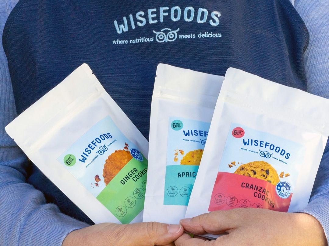 Wisefoods | Person in a navy apron holding 3 snack packs of Health Star rated cookies in Ginger, Apricot & Lemon and Cranzac