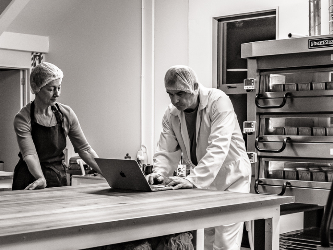 Black & white photo of Vikki & Martyn discussing the bake with laptop on wooden table and fresh loaves baking in the ovens behind