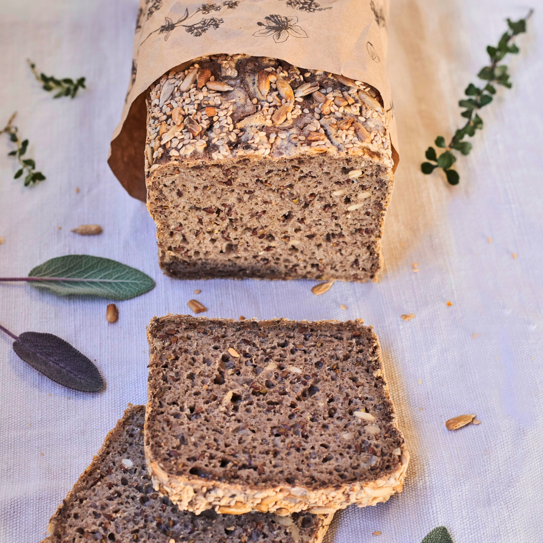 OMGoodness Sunflower &amp; Sesame &#39;Wild &amp; Free&#39; GF Organic loaf of bread on a white background with fresh herbs sprinkled alongside. Label reads Gluten Free, Paleo &amp; Vegan, Dairy, Egg &amp; Refined Sugar Free