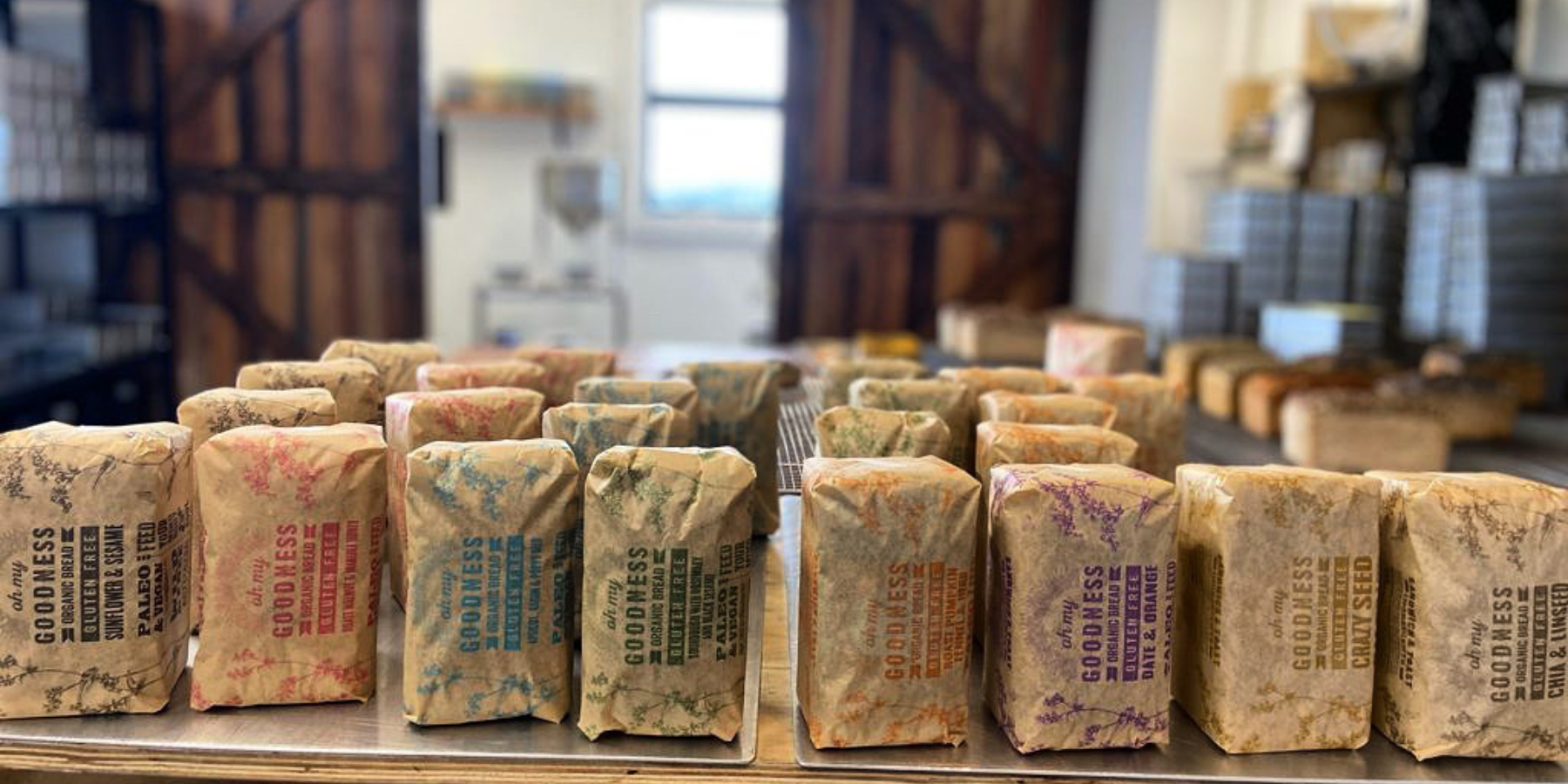 OMGoodness organic GF loaves paper-wrapped & standing on end, with the bakery prep area, wooden barn doors and baking tins out of focus in the background