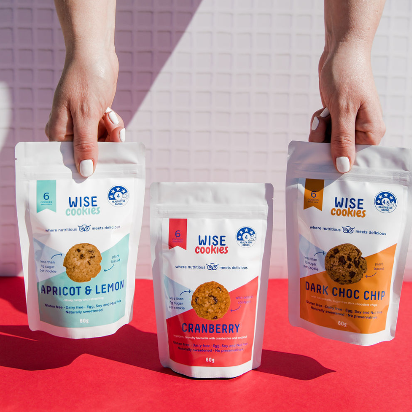 Wisefoods | Two hands lifting a Wise Cookie Apricot & Lemon and a Dark Choc Chip Snack Pack, with a Cranberry Wise Cookie snack pack in the centre on a crimson background