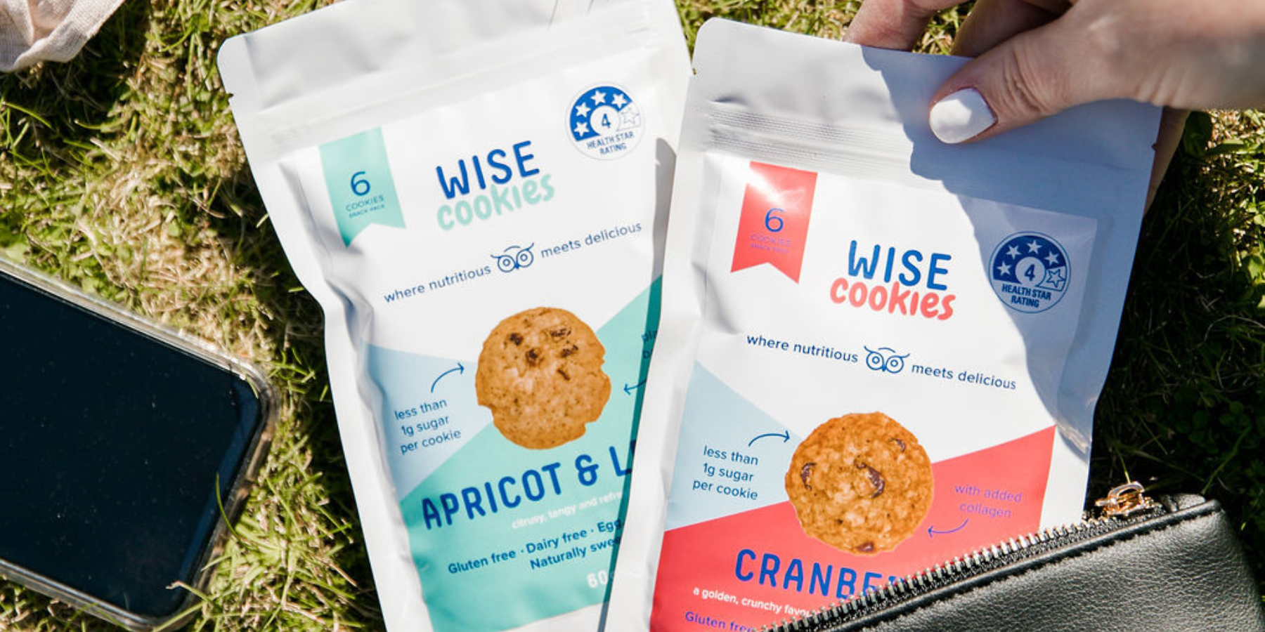 Wisefoods | Hand removing a Wise Cookie Cranberry Snack Pack from a black purse on the grass, with an Apricot Wise Cookie Snack Pack and a mobile phone in the background