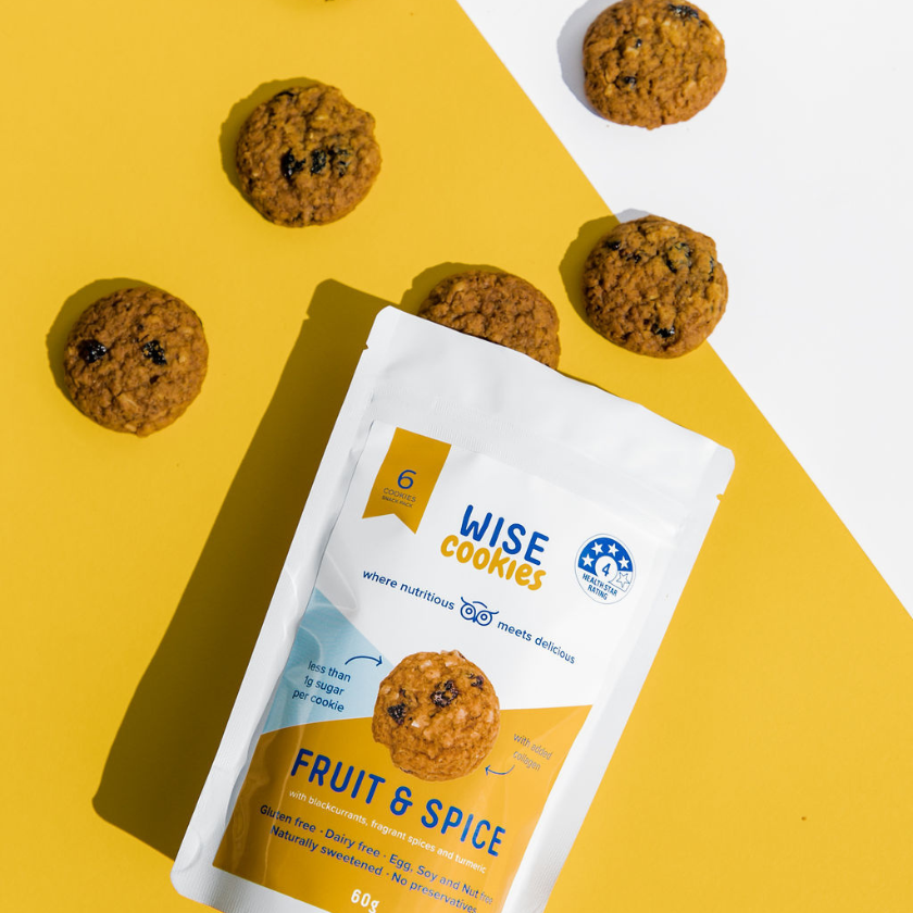 Wisefoods | Top-down view of 4.0 Health Star Rated, collagen Fruit & Spice Wise Cookies spilling out of a 60g Snack Pack on a mustard yellow background with a slash of white