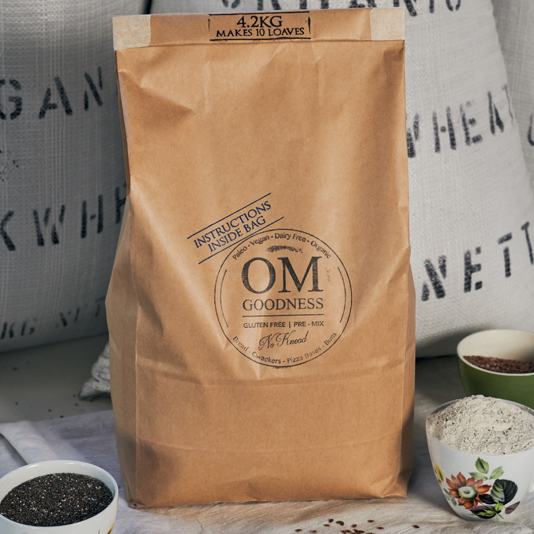 10-Loaf brown kraft bag of OMG Original Organic GF No-Knead Bread Premix standing in front of white sacks of organic buckwheat flour, with old fashioned tea cups in front filled with GF flours, chia seeds and linseeds