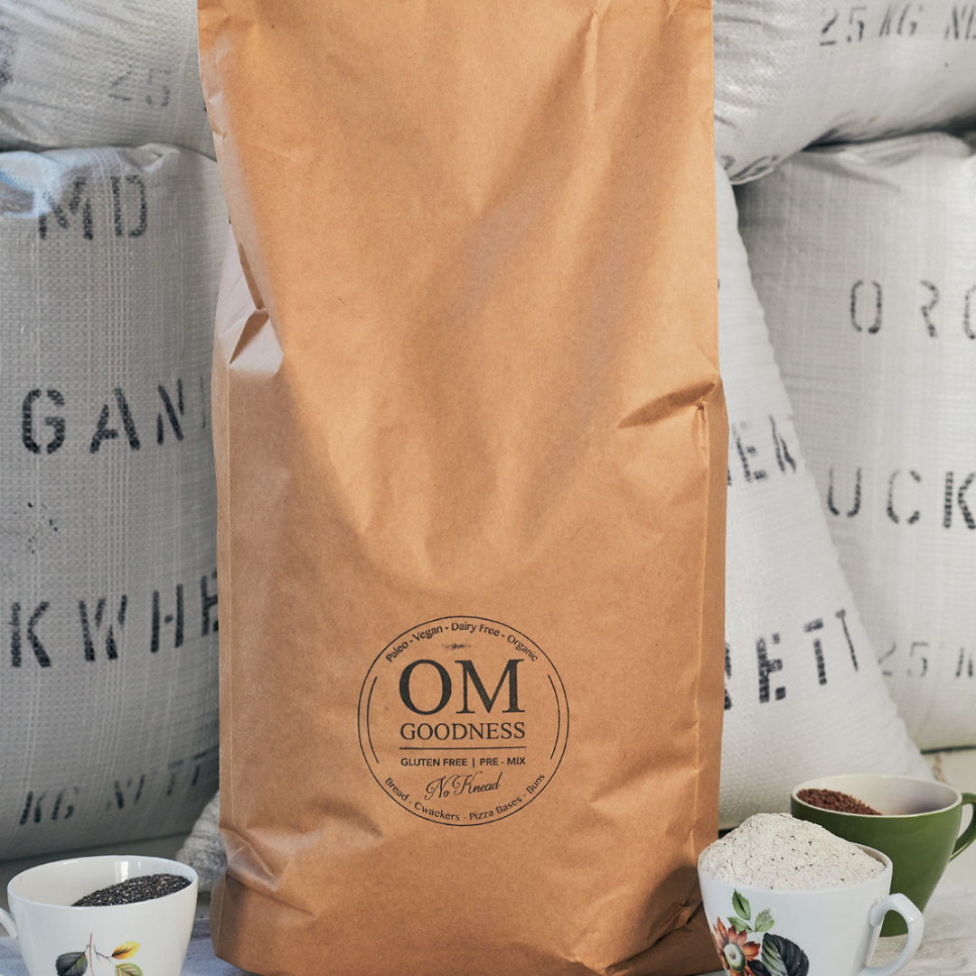 20-Loaf brown kraft bag of OMG Original Organic GF No-Knead Bread Premix standing in front of white sacks of organic buckwheat flour, with old fashioned tea cups in front filled with GF flours, chia seeds and linseeds