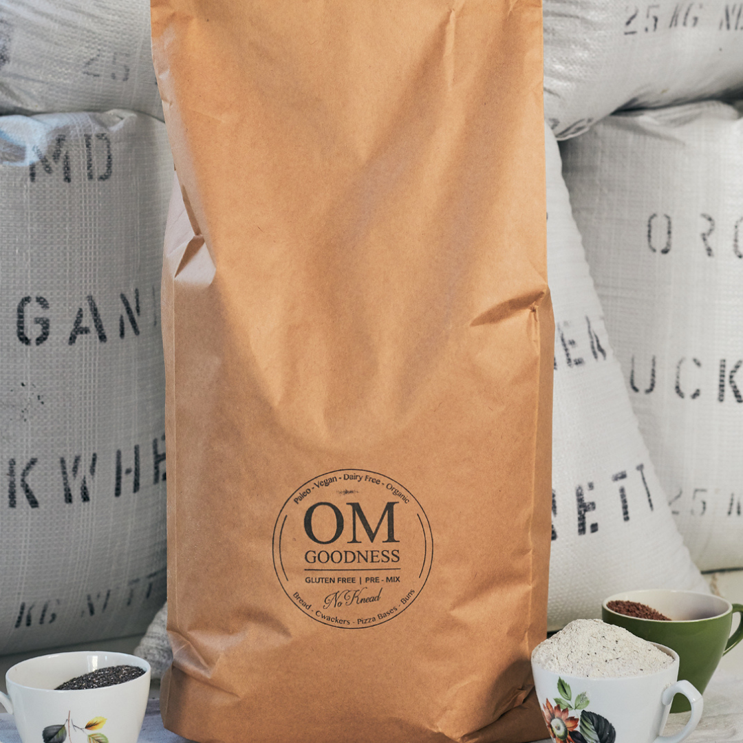 10-Loaf brown kraft bag of OMG Original Organic GF No-Knead Bread Premix standing in front of white sacks of organic buckwheat flour, with old fashioned tea cups in front filled with GF flours, chia seeds and linseeds
