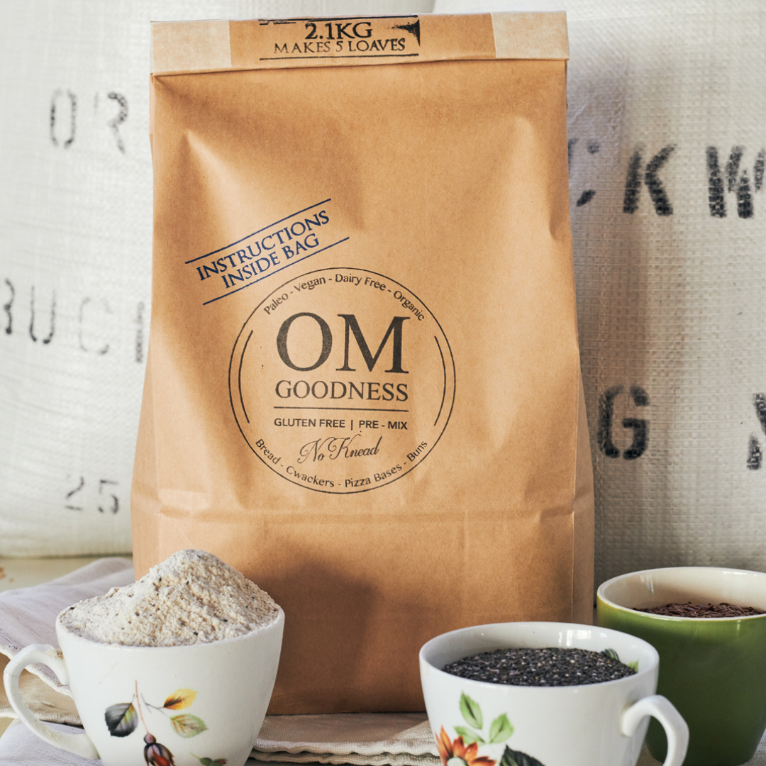2-Loaf brown kraft bag of OMG Original Organic GF No-Knead Bread Premix standing in front of white sacks of organic buckwheat flour, with old fashioned tea cups in front filled with GF flours, chia seeds and linseeds