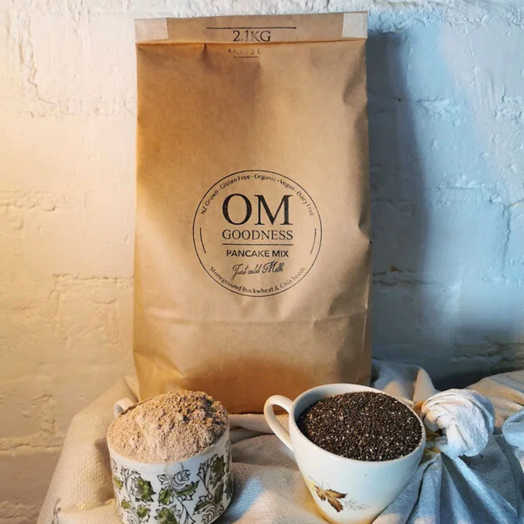 Small-size 840g brown kraft bag of OMG Organic GF Pancake Mix standing in front of white sacks of organic buckwheat flour, with old fashioned tea cups in front filled with GF flours, chia seeds and linseeds