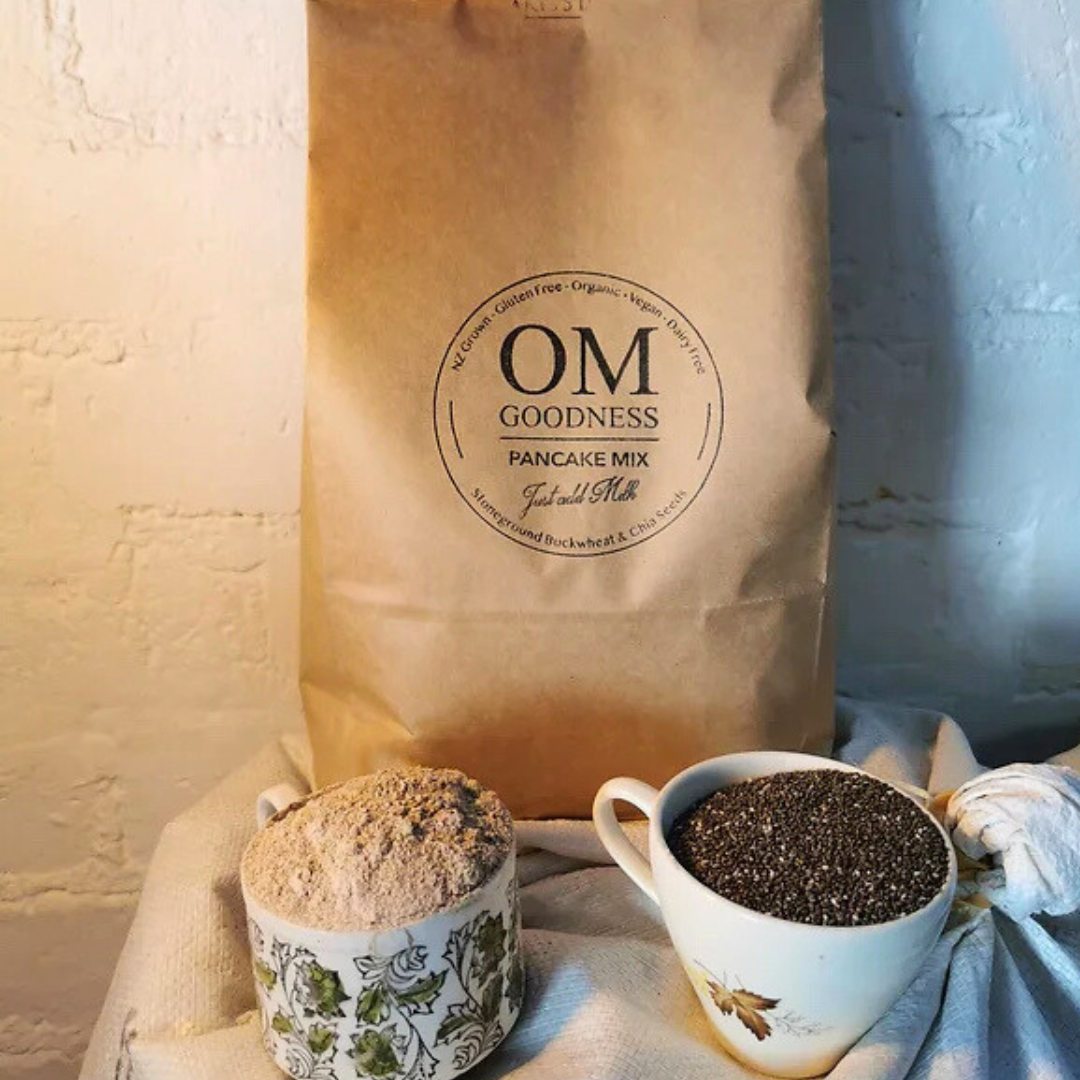 Jumbo-size 8.2 Kg brown kraft bag of OMG Organic GF Pancake Mix standing in front of white sacks of organic buckwheat flour, with old fashioned tea cups in front filled with GF flours, chia seeds and linseeds