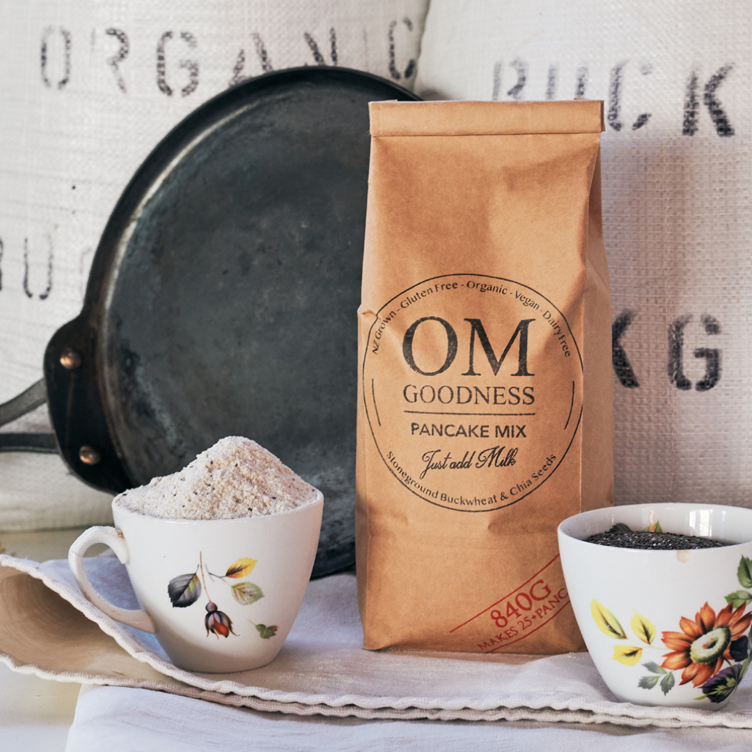 Small-size 840g brown kraft bag of OMG Organic GF Pancake Mix standing in front of white sacks of organic buckwheat flour, with old fashioned tea cups in front filled with GF flours, chia seeds and linseeds