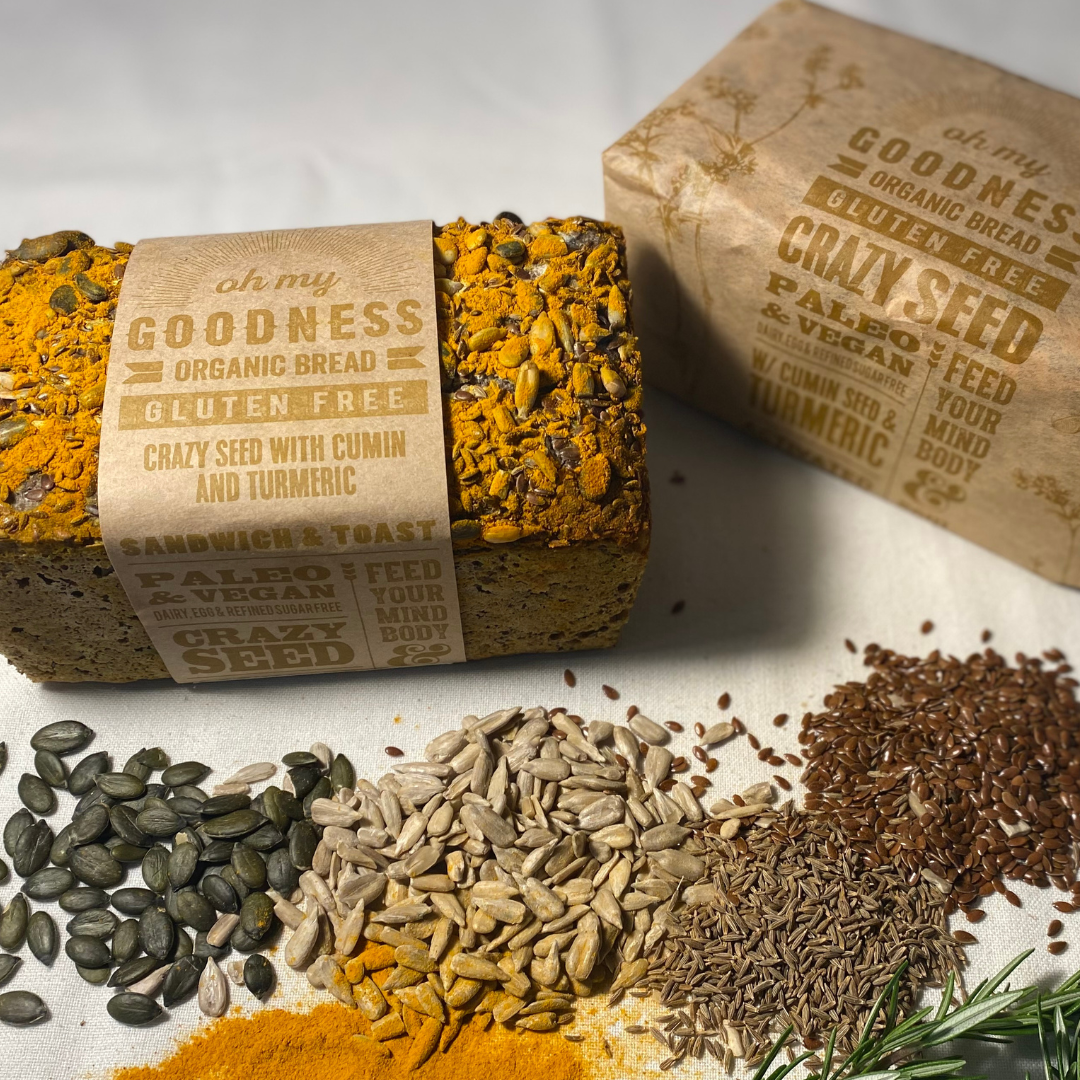 OMGoodness &#39;Crazy Seed&#39; with Cumin &amp; Turmeric GF Organic loaf of bread on a white background with pumpkin seeds, sunflower seed, cumin seeds, linseeds, turmeric powder &amp; fresh rosemary sprinkled below. Label reads Gluten Free, Paleo &amp; Vegan, Dairy, Egg &amp; Refined Sugar Free