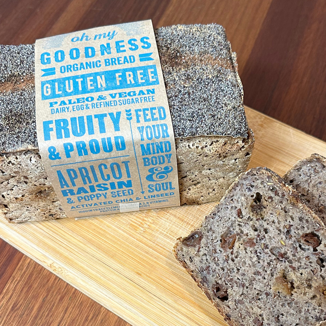 OMGoodness Apricot &amp; Raisin &#39;Fruity &amp; Proud&#39; GF Organic loaf of bread on a pale wooden board. Topped with poppy seeds &amp; cinnamon. Label reads Gluten Free, Paleo &amp; Vegan, Dairy, Egg &amp; Refined Sugar Free
