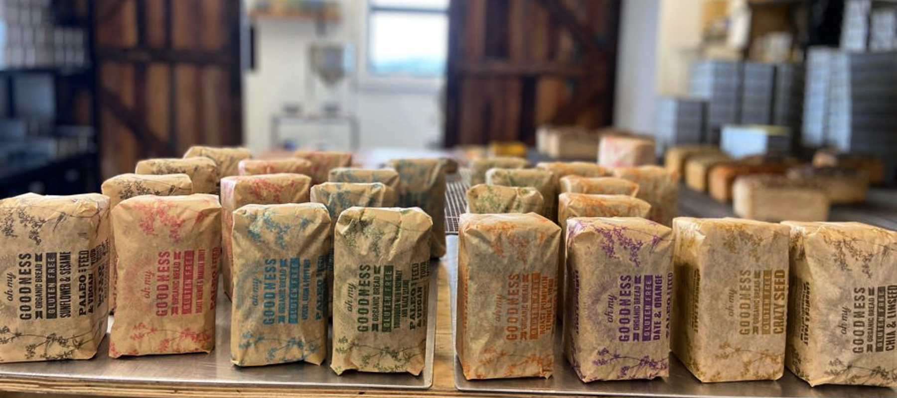 OMGoodness bread wrapped & standing on end with the bakery prep room wooden doors and stacks of bread tins out of focus in the background