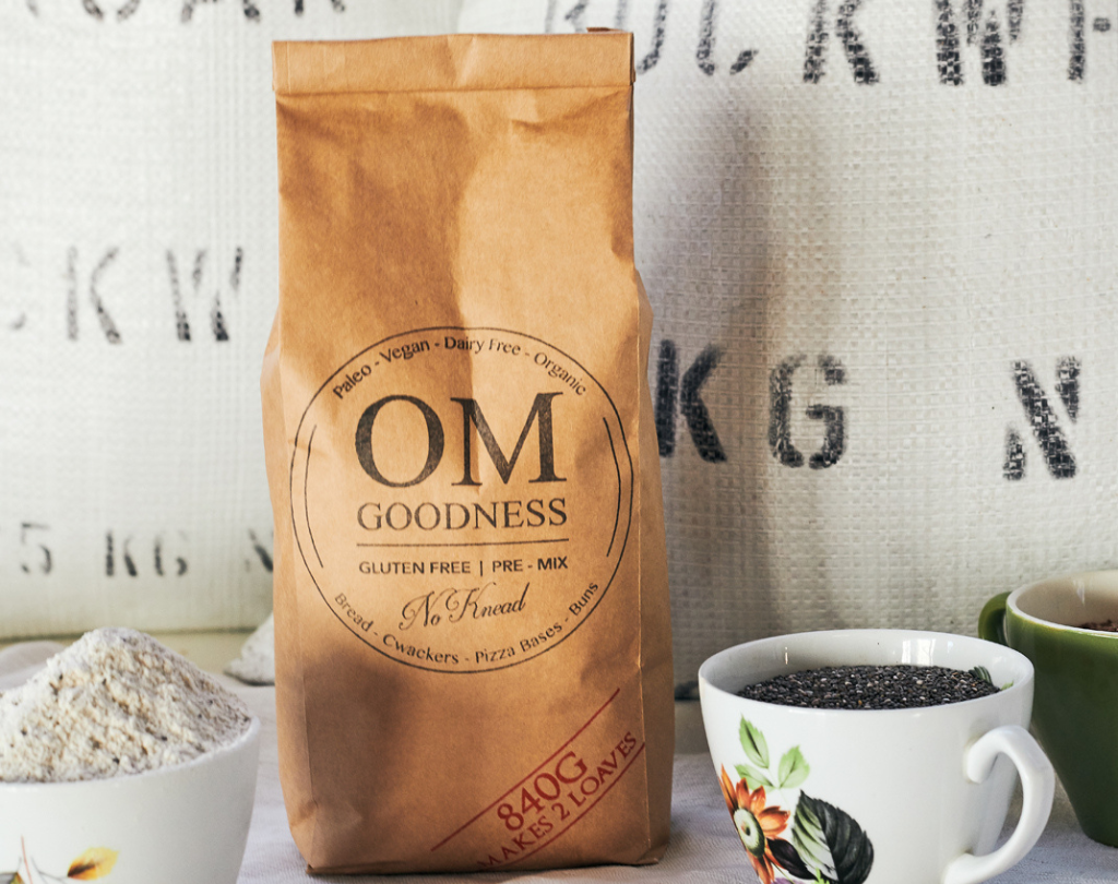 Pack of OMGoodness 2-loaf organic & gluten free no-knead bread premix with tea cups containing tapioca, chia & lindseeds & organic buckwheat sacks in the background 