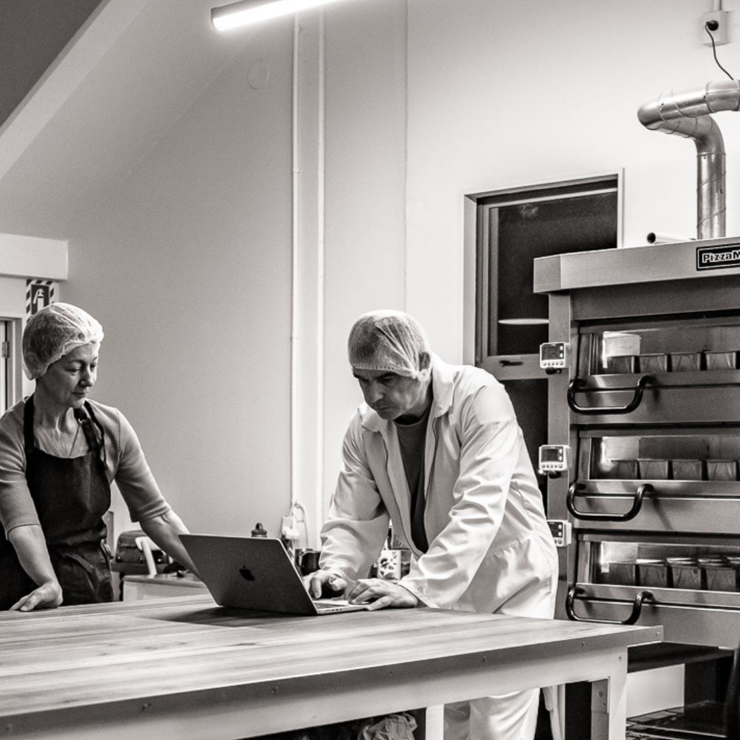 Black & white image of Vikki & Martyn planning the bake on a laptop with fresh OMGoodness loaves baking in the oven behind