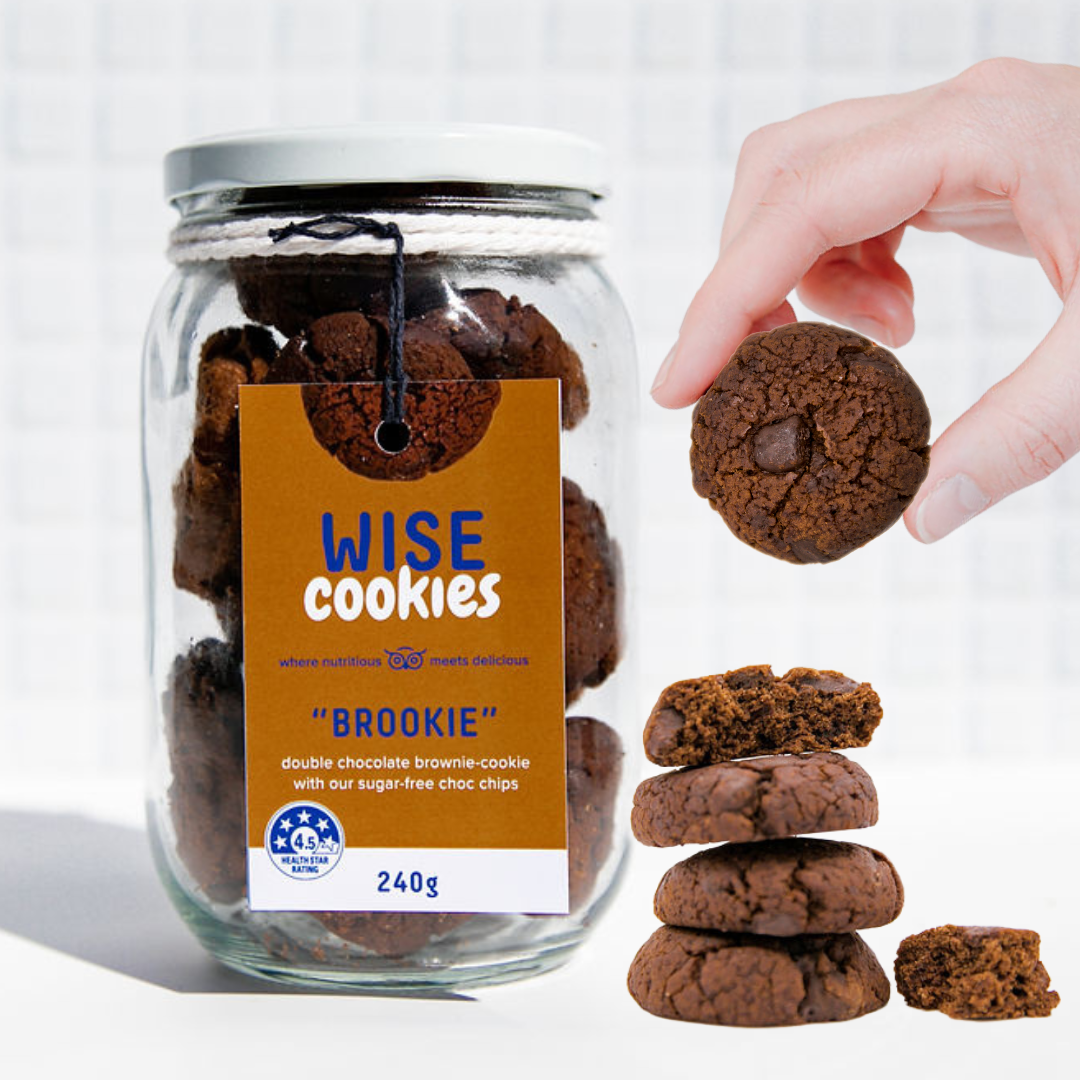 Wisefoods | 4.5 Health Star rated, allergy-friendly Double Choc &quot;Brookie&quot; Wise Cookies in a glass jar with a stack of loose cookies and a hand holding one between thumb and forefinger to the right of the jar
