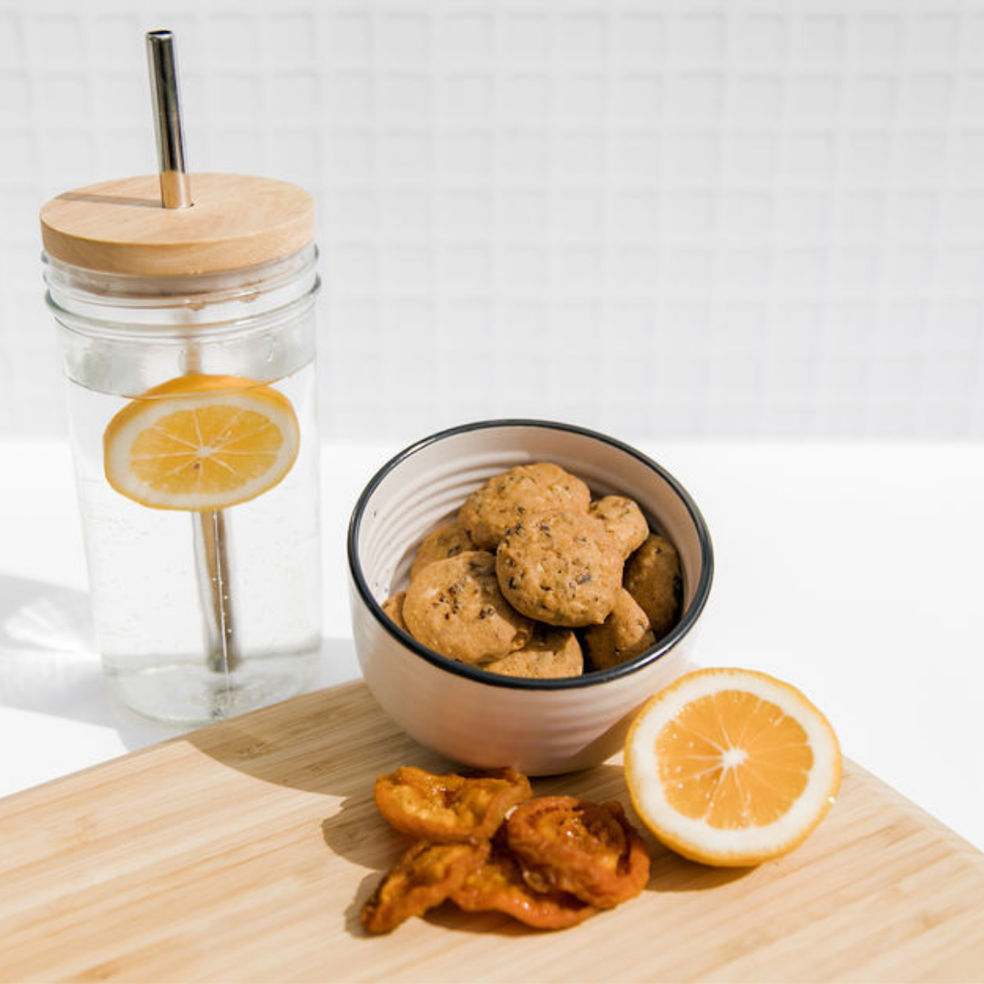 Wisefoods | Apricot and Lemon 4.0 Health Star rated, allergy-friendly, plant based Wise Cookies in a pottery dish on a timber board. With dried apricots and fresh cut lemon alongside and a sipper glass of water with a slice of lemon in the background