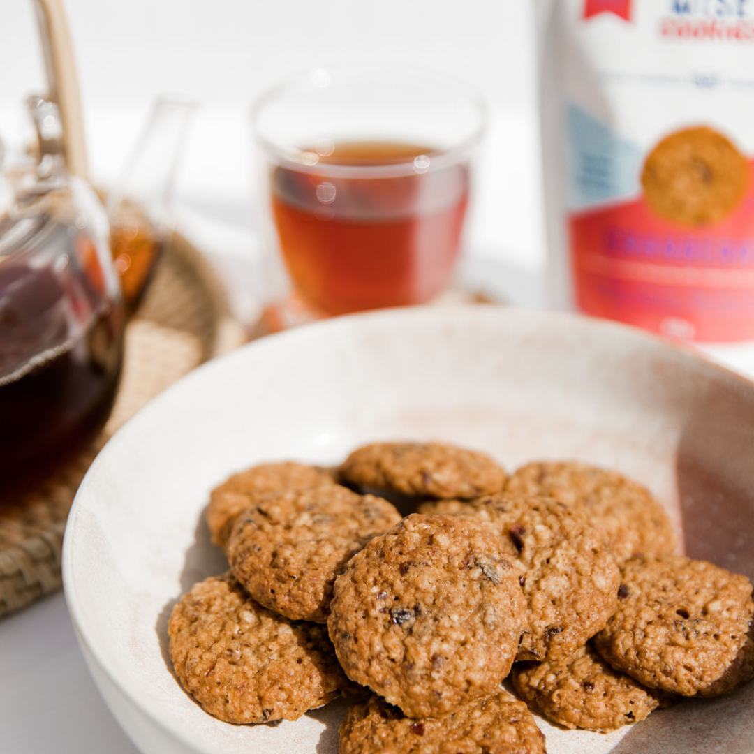 Wisefoods | Close up of a white plateful of 4.0 Health Star rated, allergy-friendly, collagen Cranberry Wise Cookies, with a thermo-glass of black tea, glass teapot out of focus in the background 