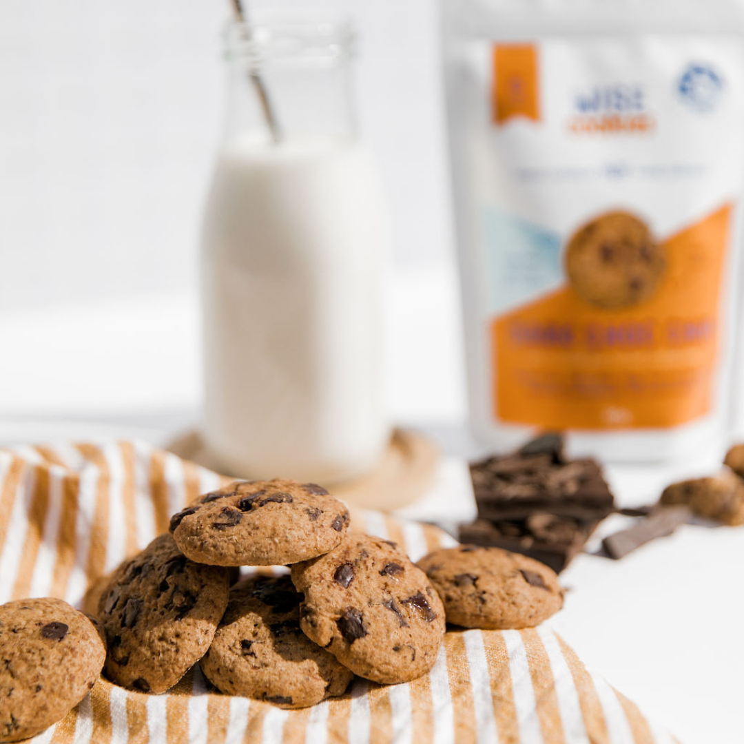 Wisefoods | Dark Choc Chip 4.5 Health Star rated, allergy-friendly and plant based cookies on a striped orange cloth with a bottle of oat milk, choc pieces and a Snack Pack in the background