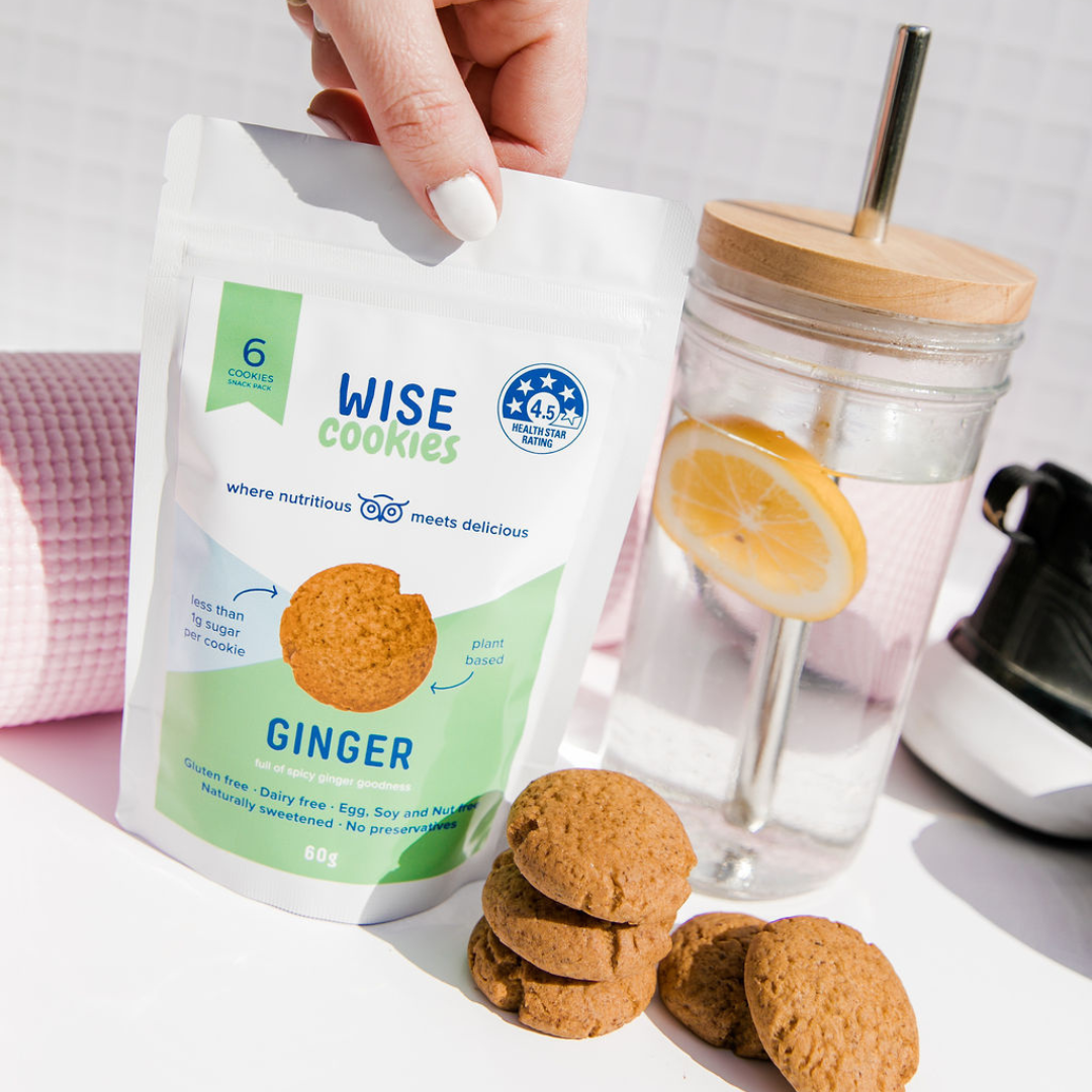 Wisefoods | Hand holding a Snack Pack of 4.5 Health Star, allergy-friendly, plant based Ginger Wise Cookies, displayed with a stack of Ginger cookies and a sipping glass of lemon water with a pink yoga mat and the edge of a black sports shoe in the background